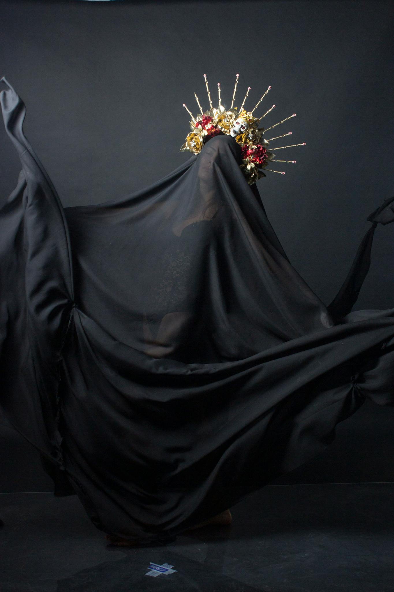 A model dances with black fabric, adorned with a floral headdress in a moody studio setting.